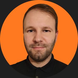 mężczyzna koło 30 w czarnej koszuli z brodą na pomarańczowym tle projektant stron internetowych Mariusz Przyimka webnim uk